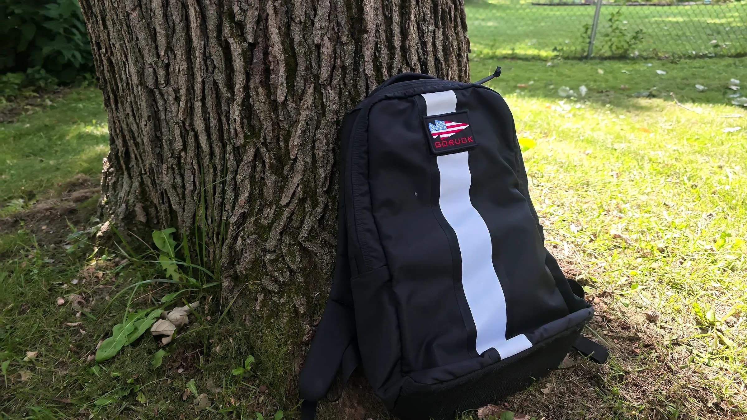 Black GORUCK Basic Rucker with a white reflective stripe and flag patch, leaning against an oak trunk on summer grass