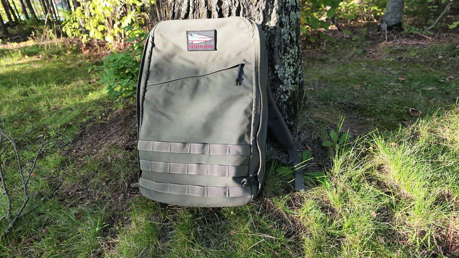 A ranger green GORUCK GR1 leaning against a tree on a grassy trail edge