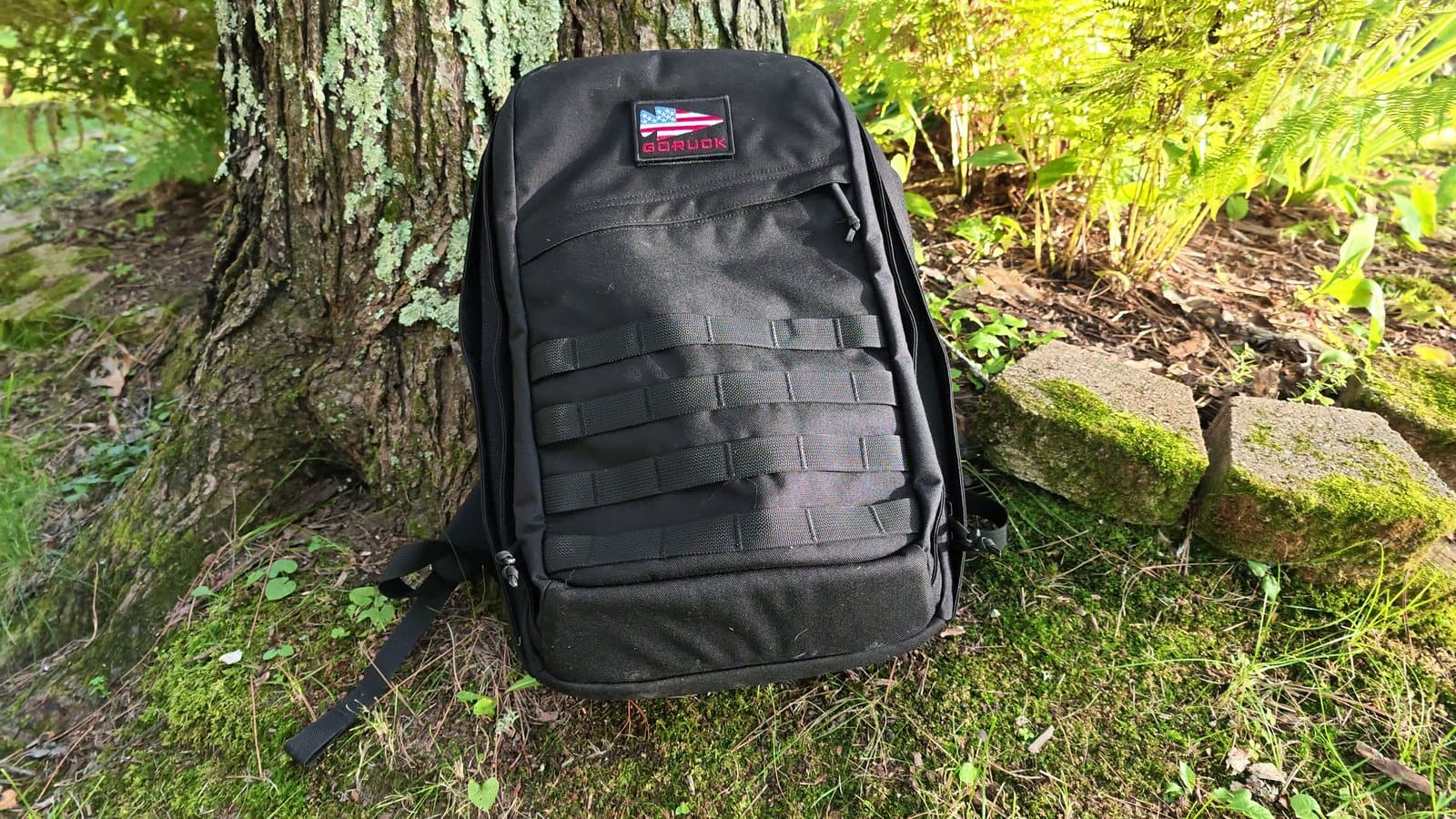 The black GR2 34L standing against a tree, front MOLLE rows and flag patch facing the camera