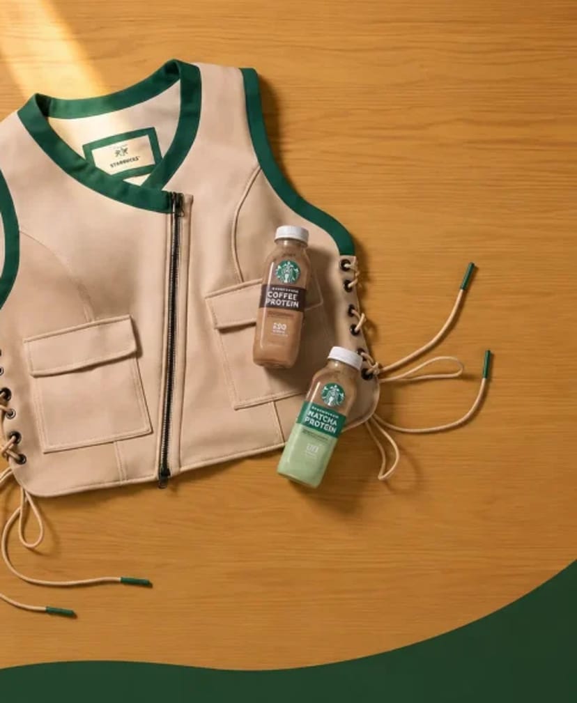 Close-up flat-lay of the Starbucks weighted vest with sage green binding, lace-up sides, and two Starbucks Coffee & Protein bottles on a warm wood surface