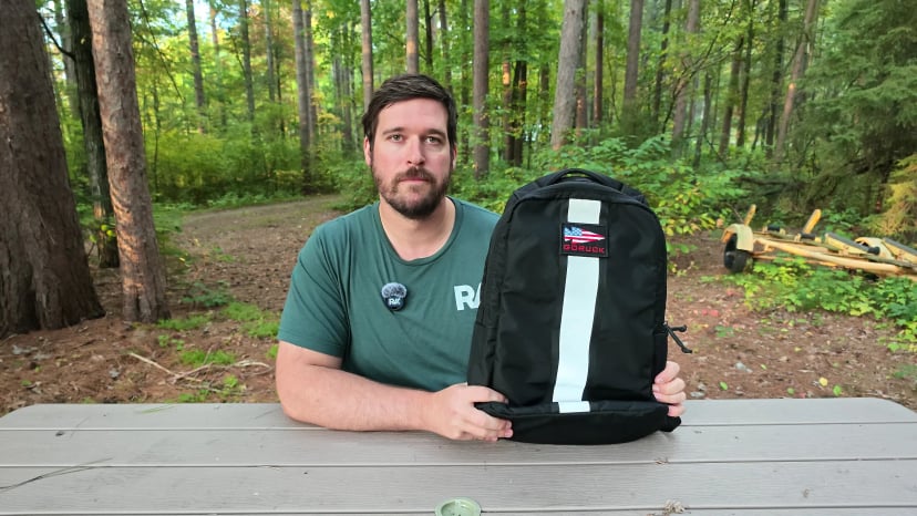 Joel Kelly at a picnic table in the woods holding up the black 17L GORUCK Basic Rucker
