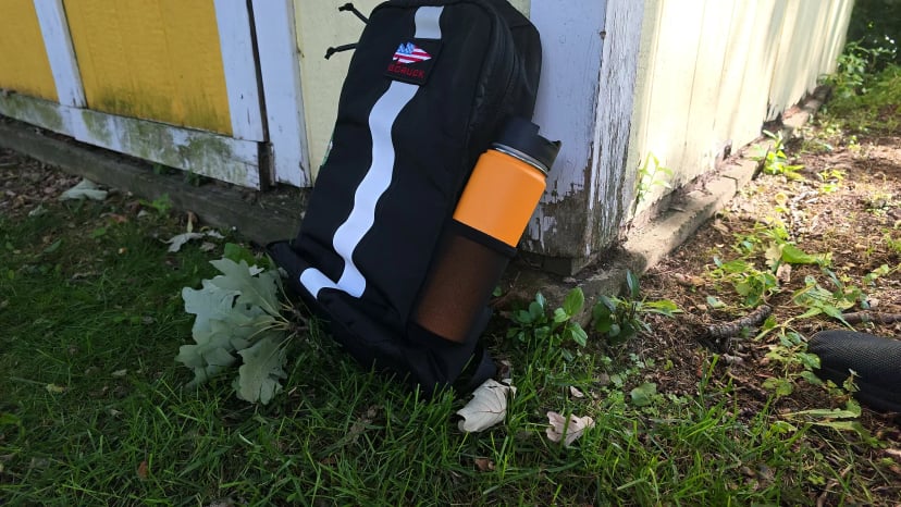 Basic Rucker propped against a yellow shed with an insulated bottle in its elastic side pocket
