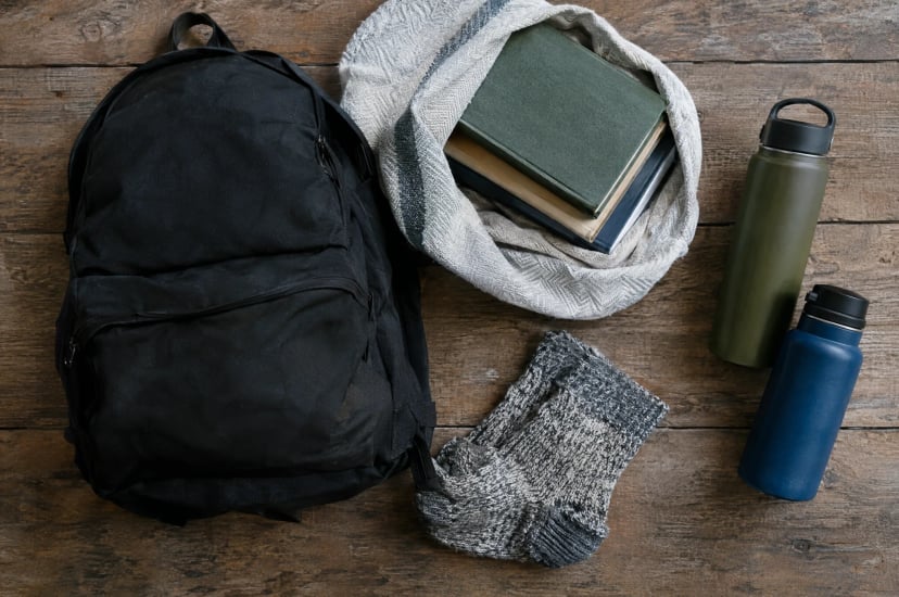 A plain black daypack on a wooden table beside hardcover books wrapped in a towel, two water bottles and wool socks